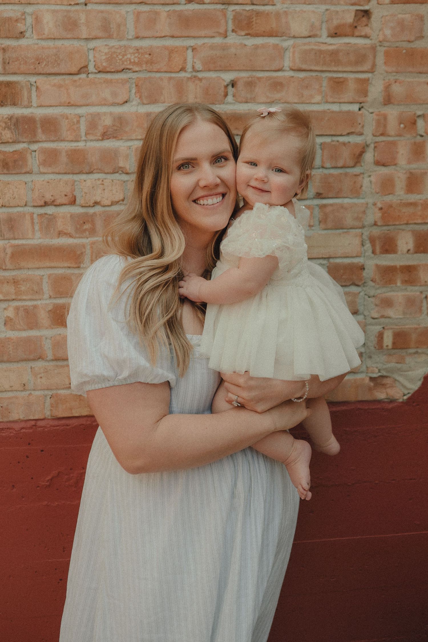 A happy blonde mother in a blue dress holds her smiling baby girl against a brick wall.