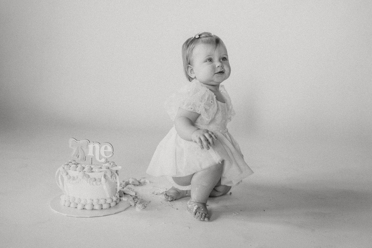 Black and white photo of baby girl standing beside her first birthday smash cake.