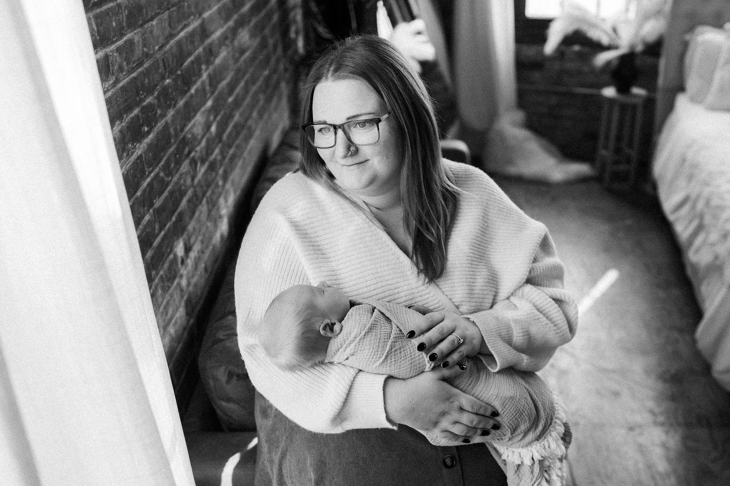 Black and white photo of a woman wearing glasses holding a newborn baby indoors near a brick wall.