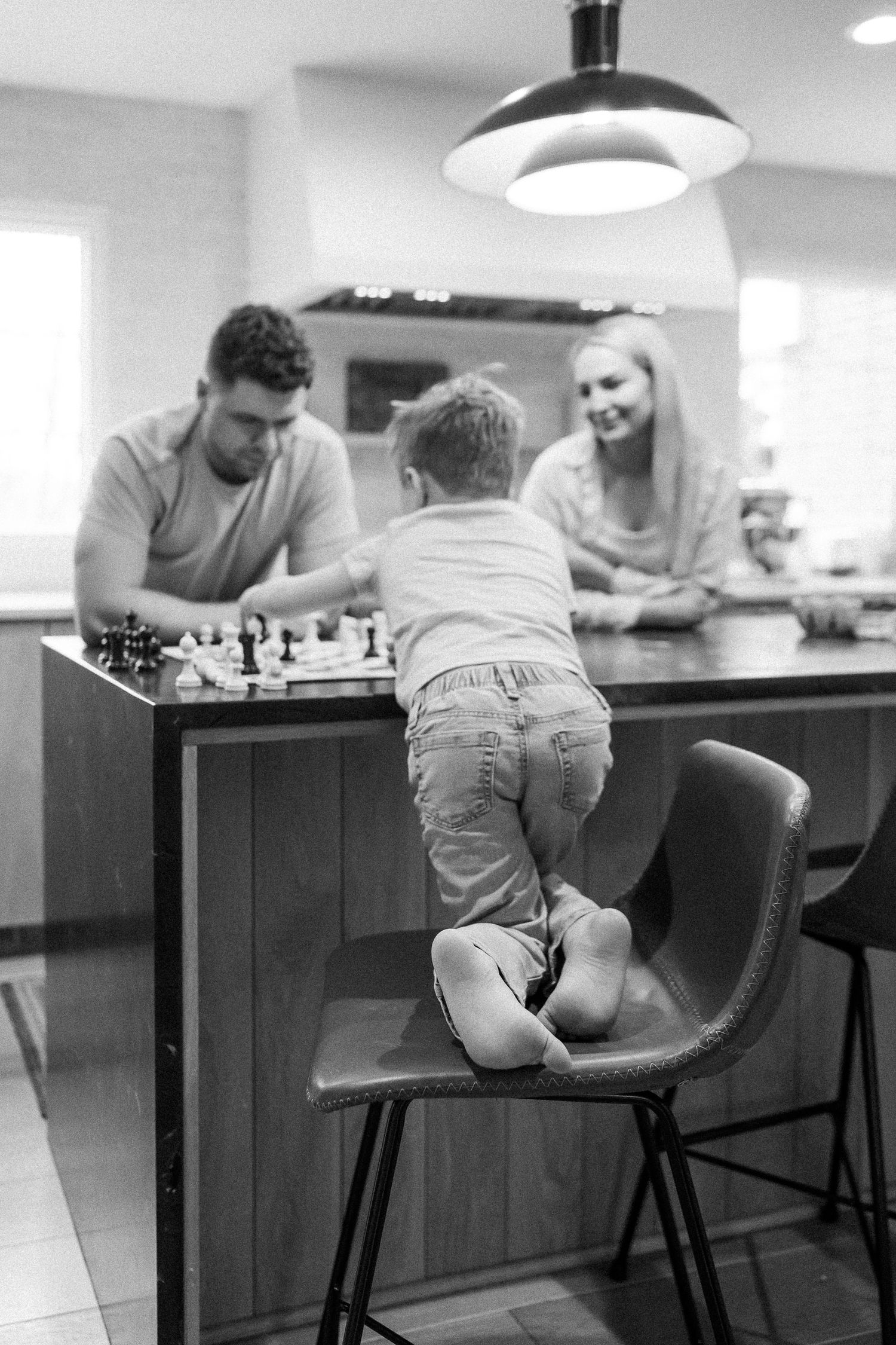 A young child stands on a chair watching parents play chess at a kitchen island in black and white.