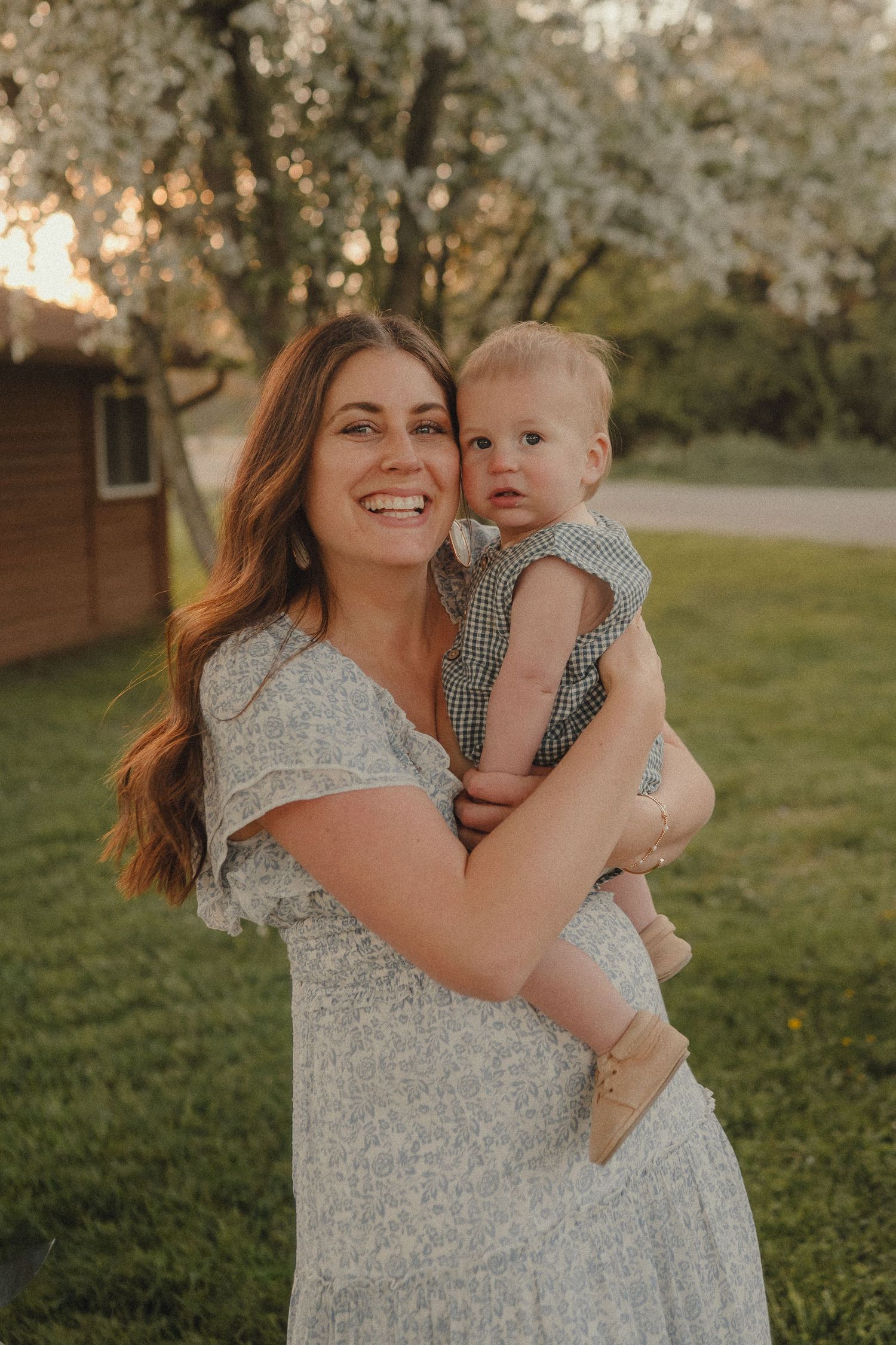 A smiling mother in a floral dress holds her baby boy outdoors near blooming trees at sunset.