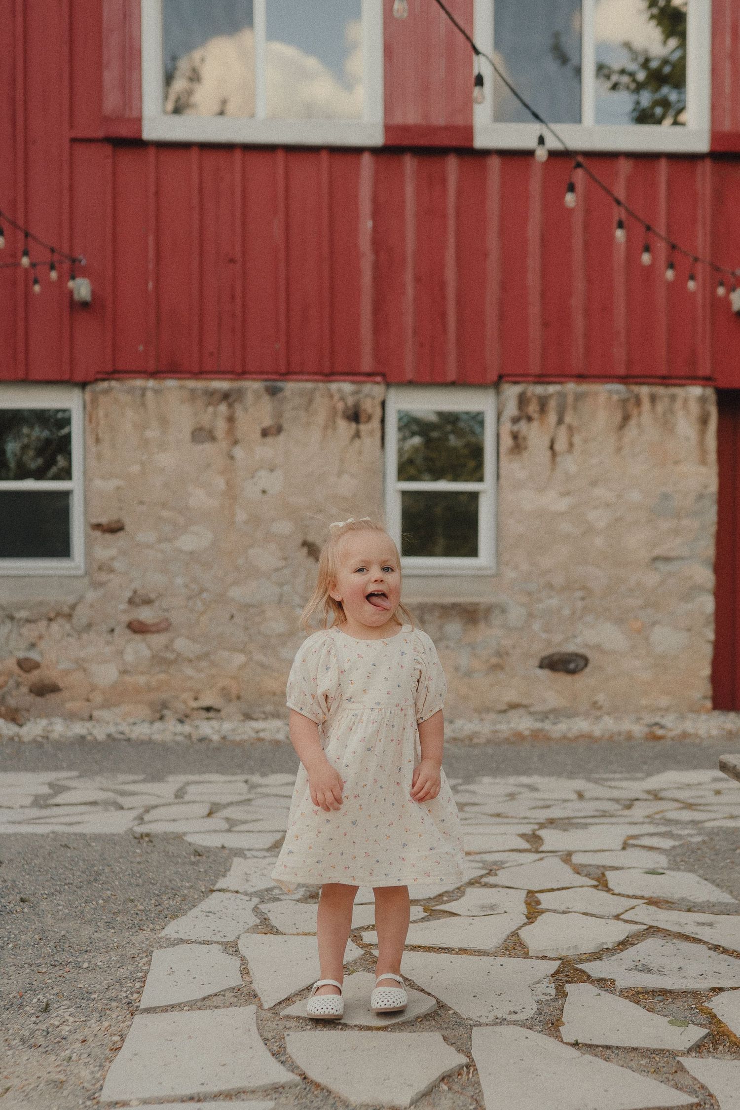 Adorable toddler girl in cream dress laughing outdoors near a rustic red barn on stone patio.