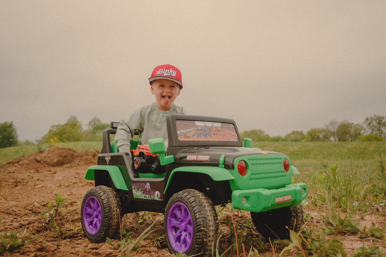 Young boy in red cap driving green and purple toy Jeep on dirt field outdoors.