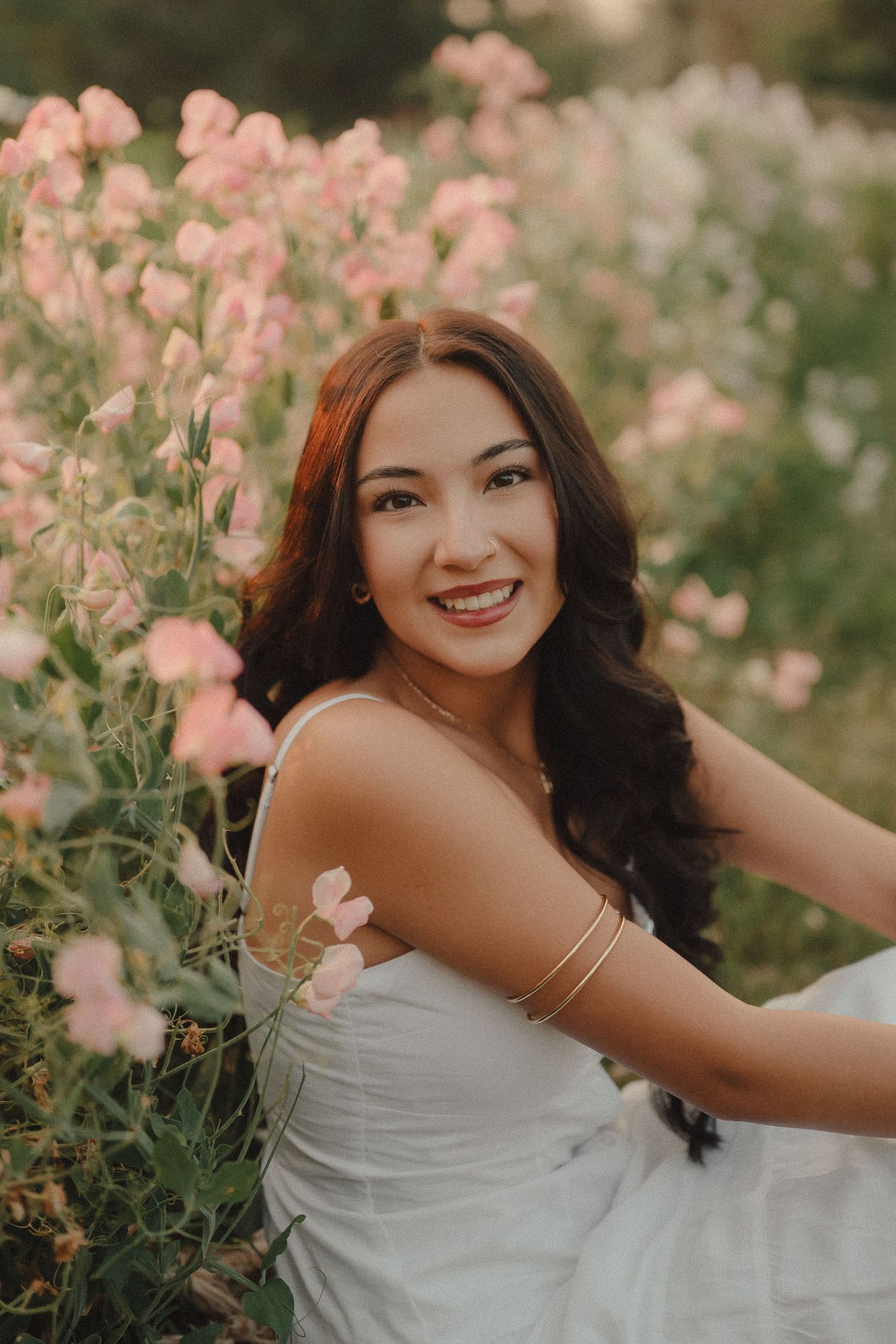 Smiling young woman in white dress surrounded by pink wildflowers at sunset.
