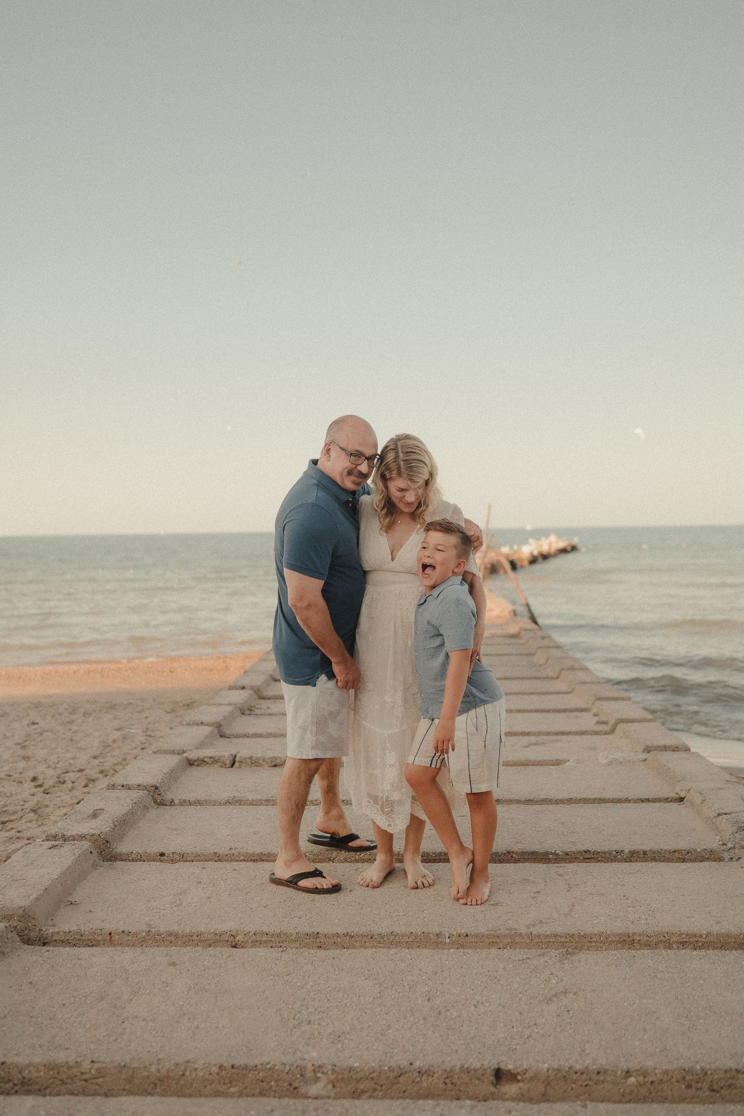 Family of three laughing together on a beachside pier during a warm sunset evening.