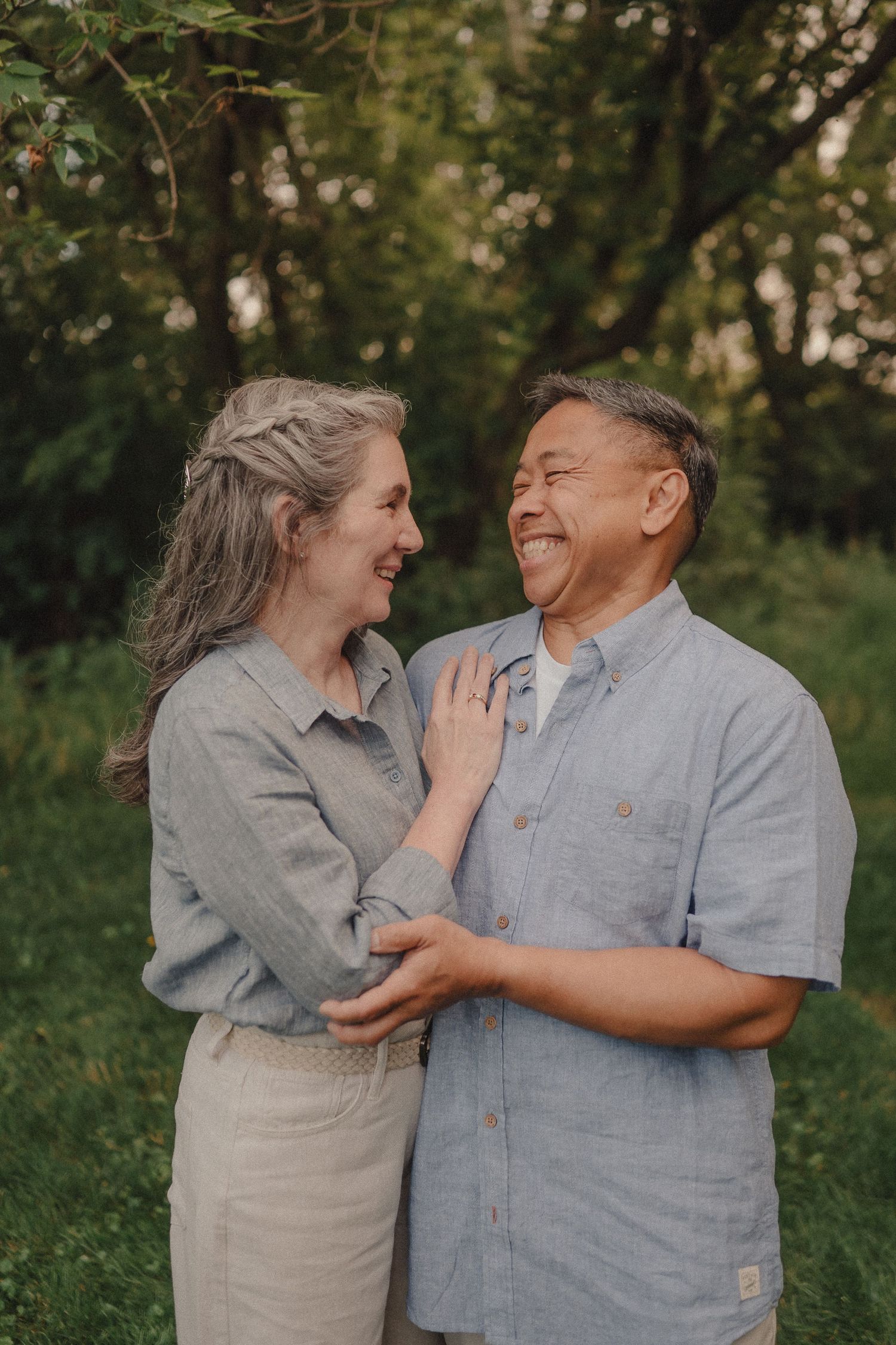 Laughing middle-aged interracial couple embracing outdoors in lush green park setting.