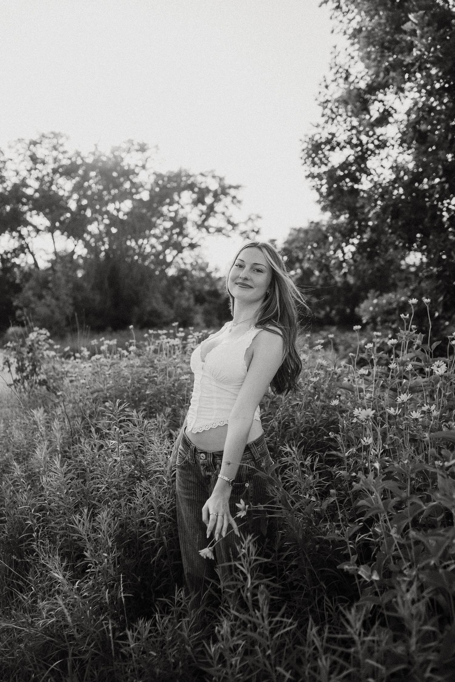 Young woman in white top and jeans standing in wildflower field, black and white portrait photography.
