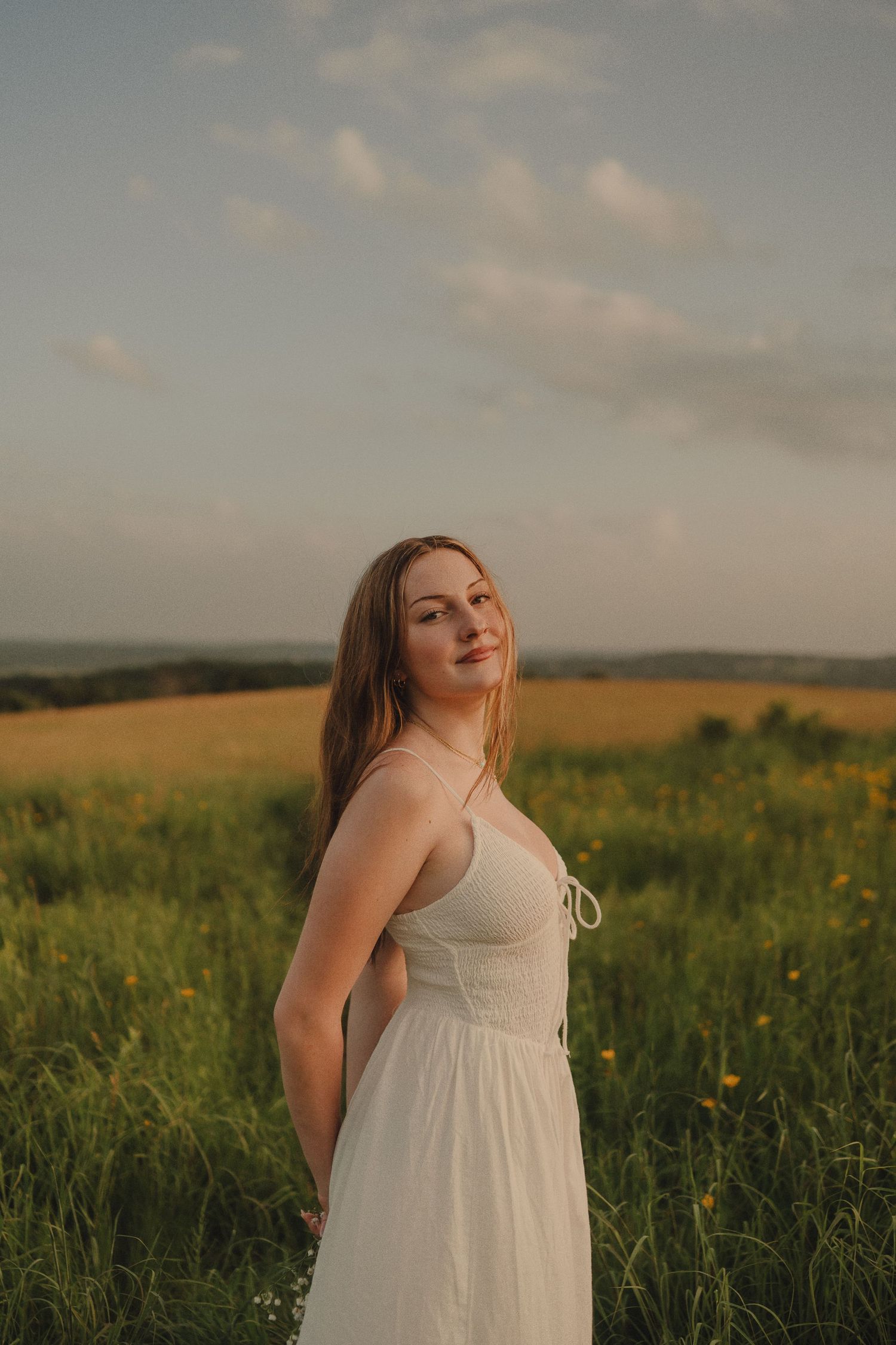 Young woman in white maxi dress standing in a green meadow under a partly cloudy sky at sunset.