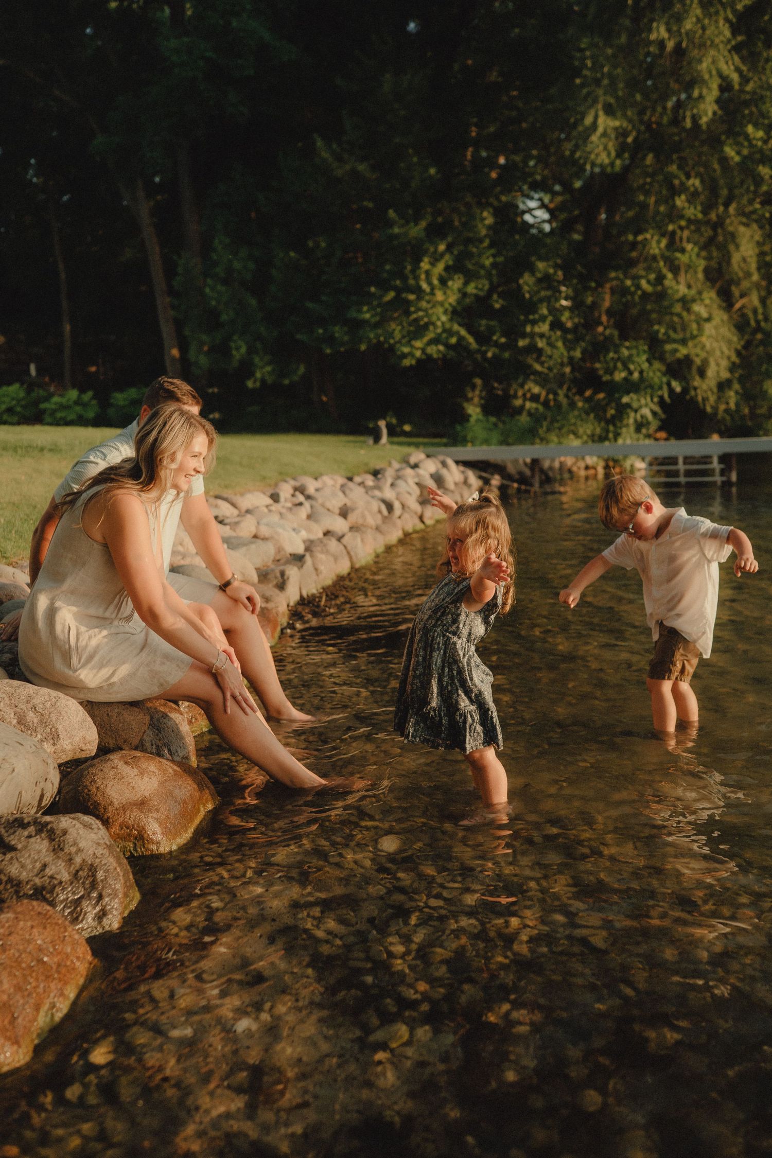 A mother sits on rocks watching her two young children play in shallow water during golden hour sunset.