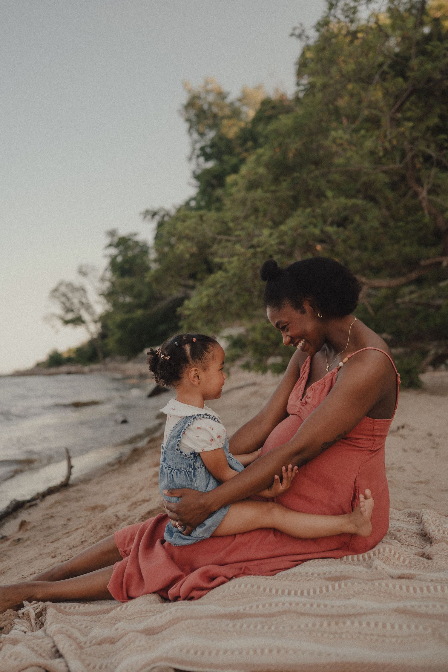 Pregnant mother in coral dress sitting on blanket on beach, sharing tender moment with young daughter.