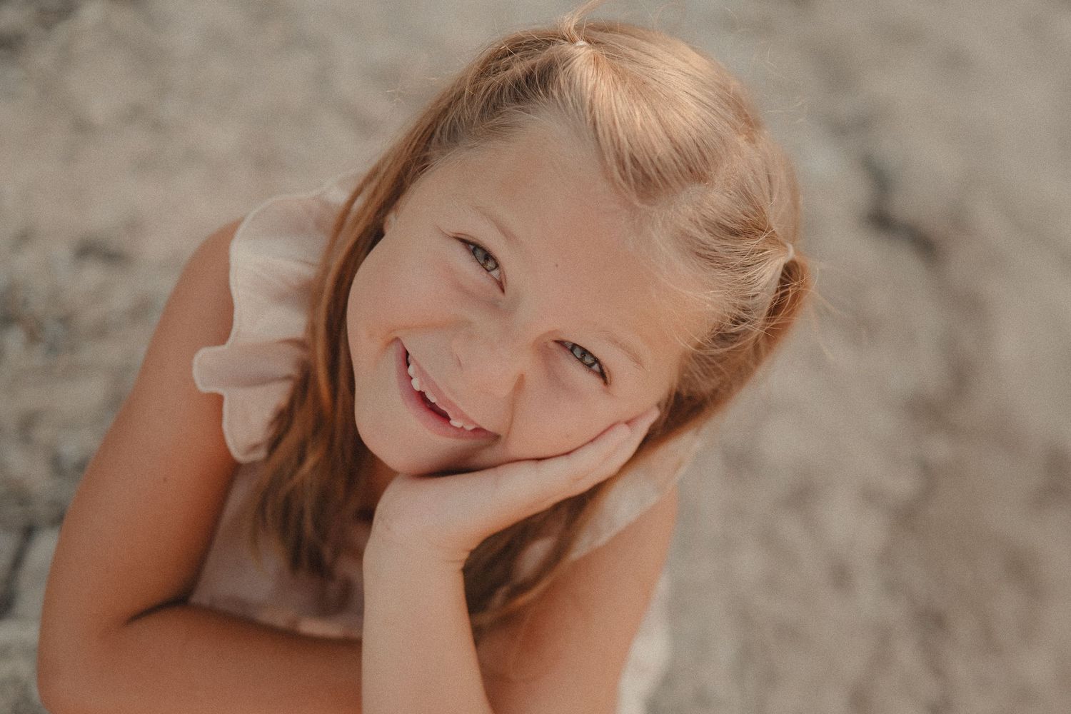 Smiling young girl with brown hair resting chin on hand, photographed outdoors in warm natural light.