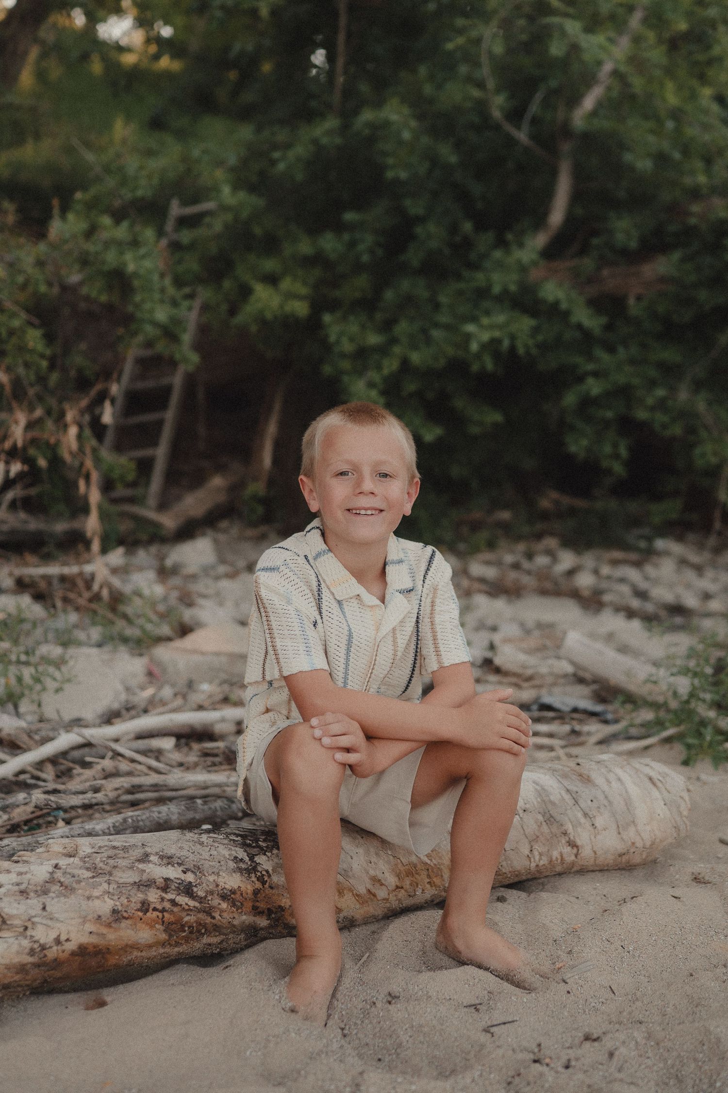Smiling young boy sits on driftwood log on a sandy beach with trees behind him.