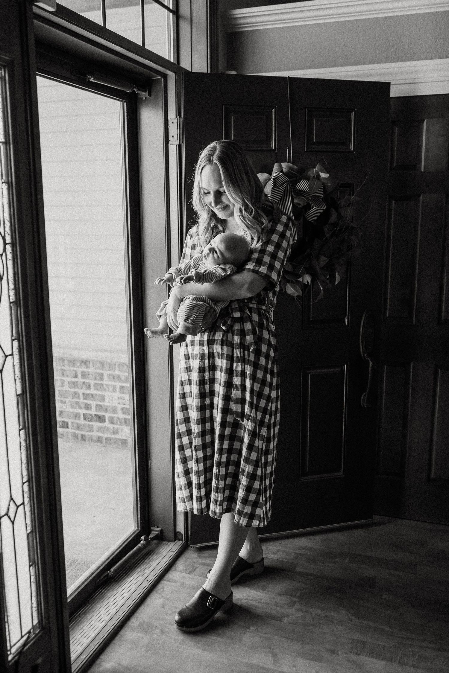 Young blonde mother in plaid dress cradling newborn baby by front door in black and white photo.