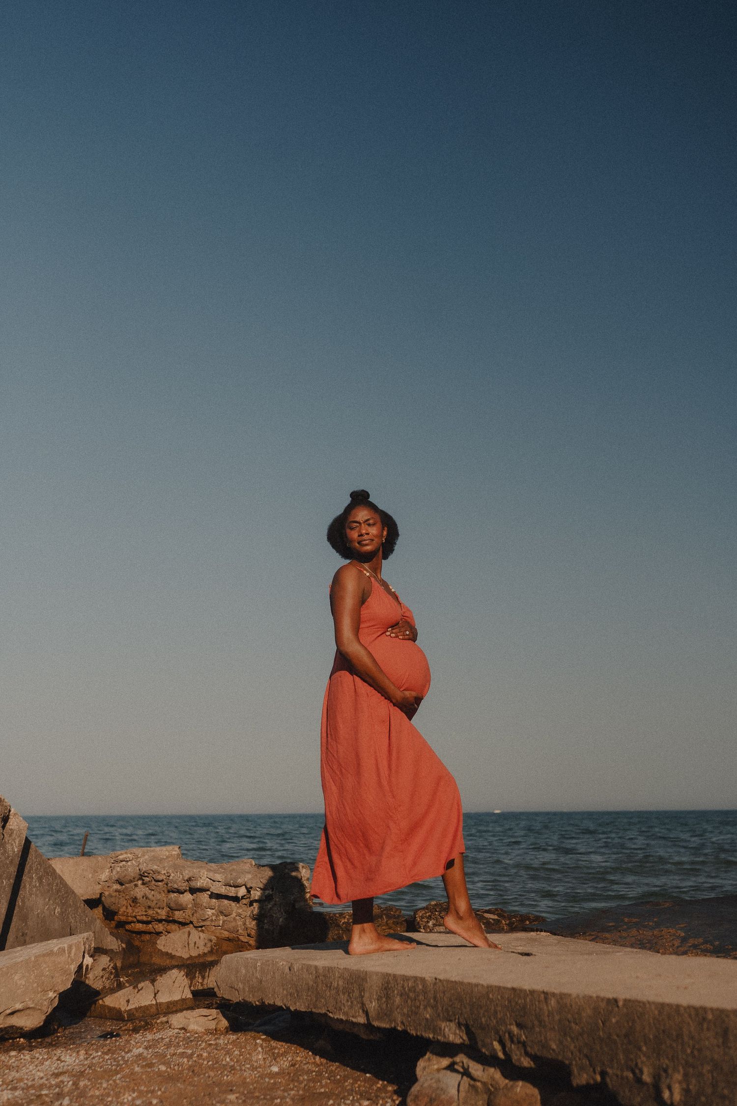 Pregnant woman in coral dress and hat stands on rocks by the ocean under a clear blue sky.