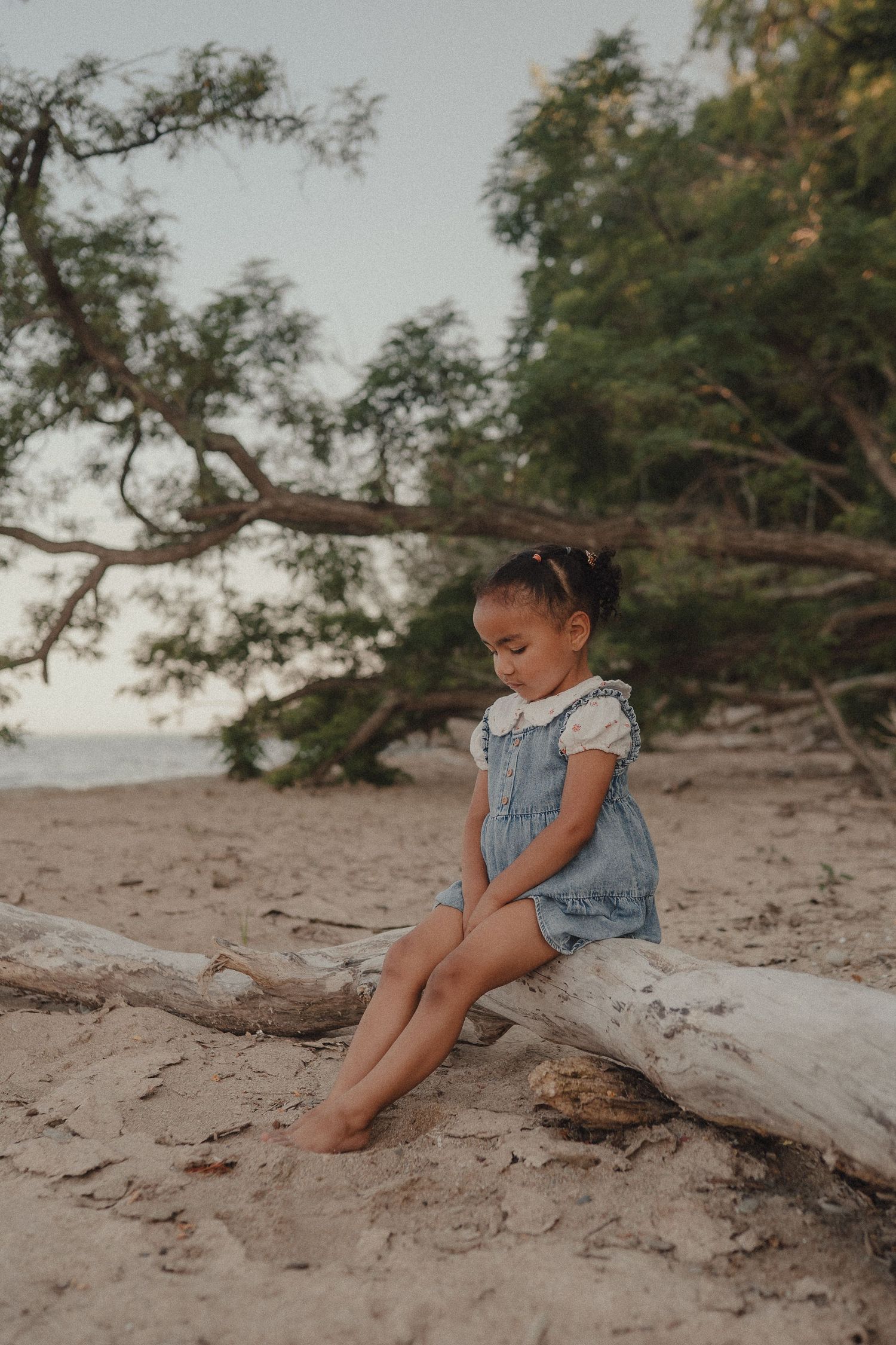 Young girl in denim romper sits barefoot on driftwood log at sandy beach with green trees backdrop.
