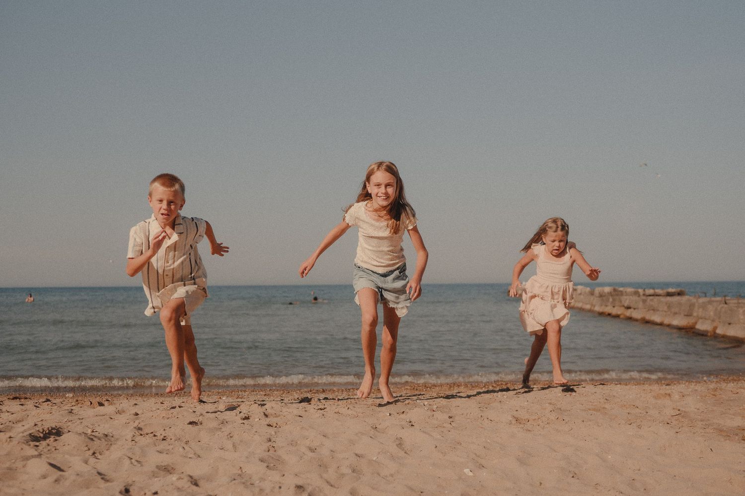 Three happy children running barefoot on a sandy beach with calm ocean water in the background on a sunny day.