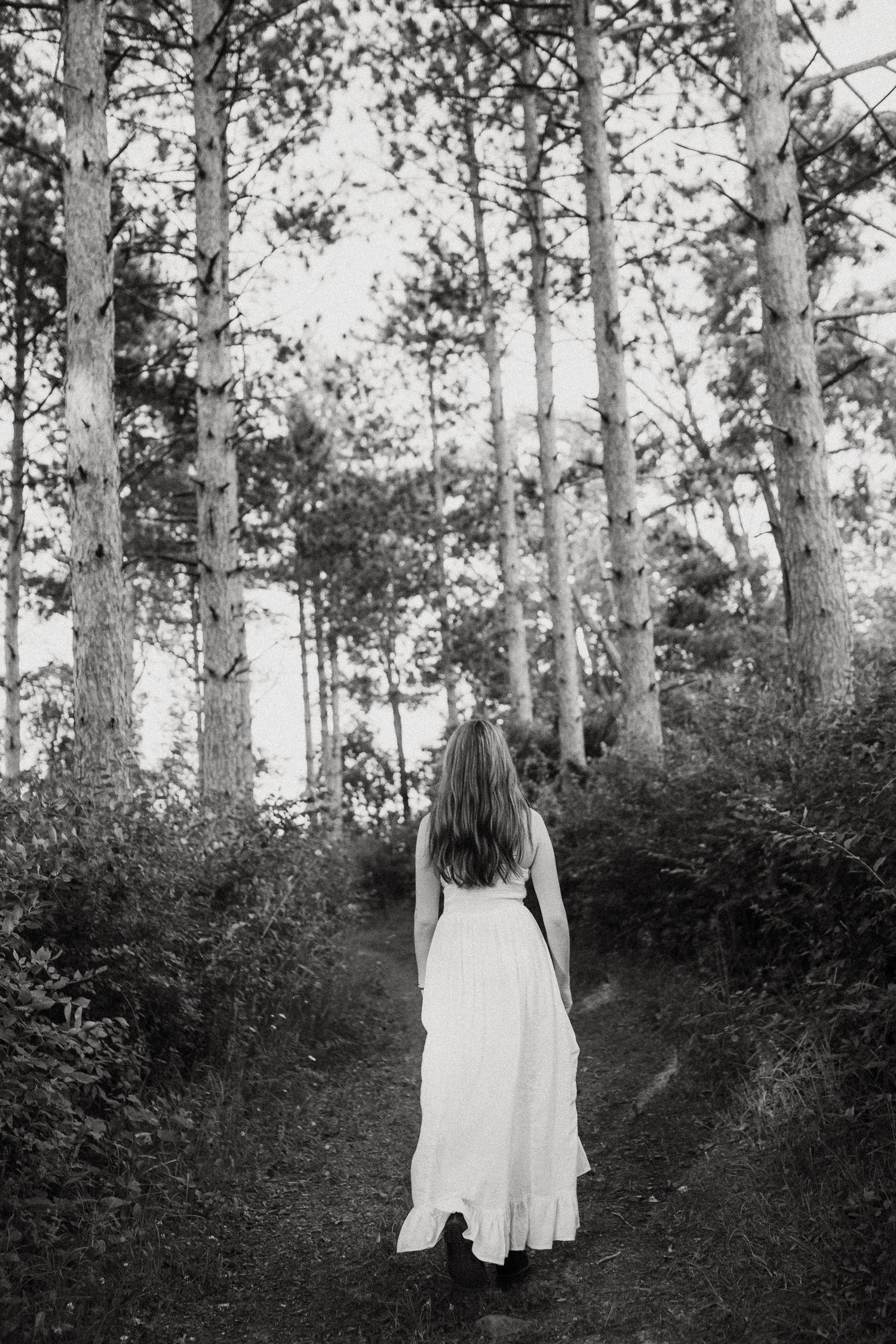 Black and white photo of a woman in a white dress walking along a serene forest path among tall trees.