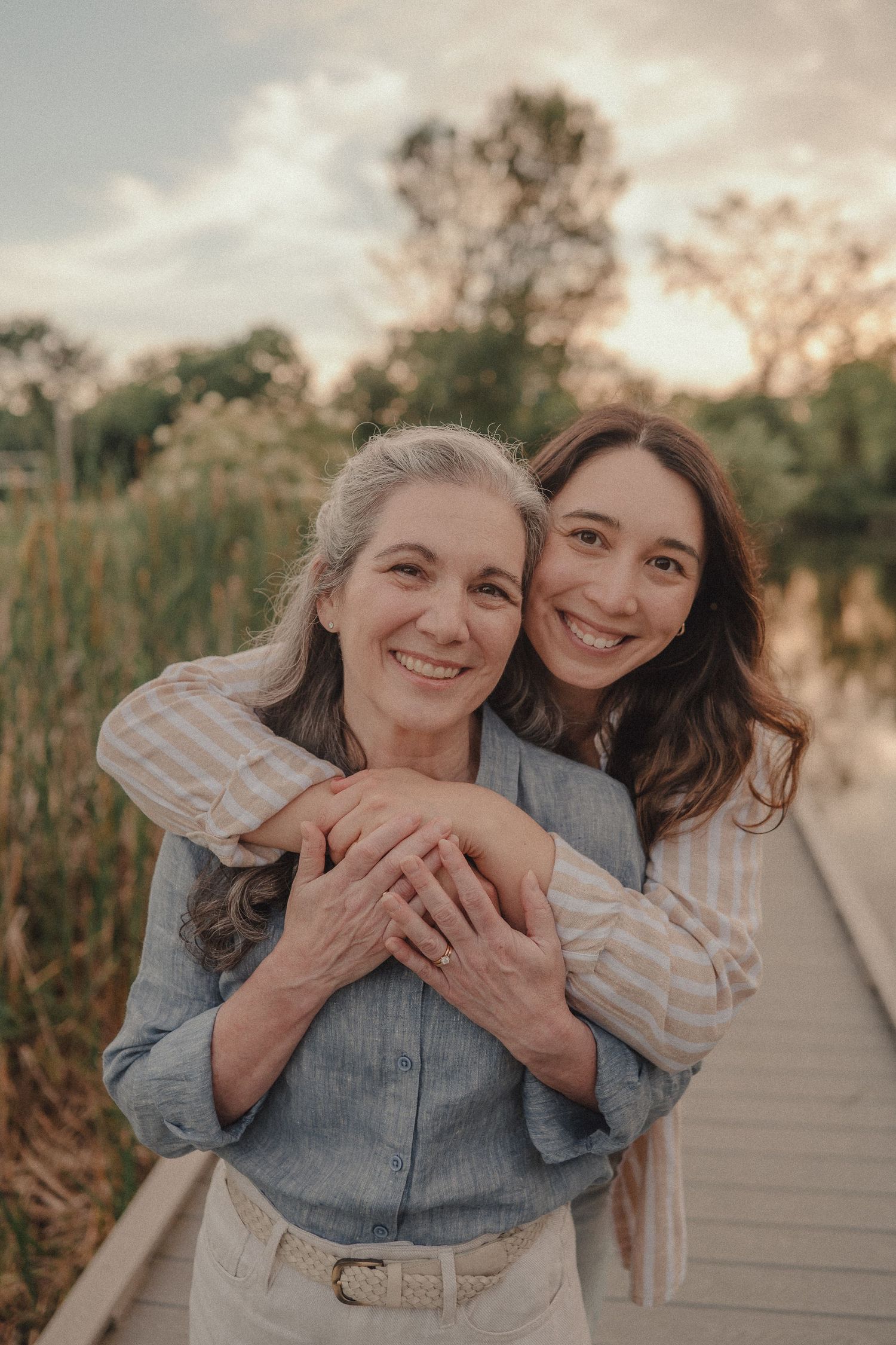 A young woman hugging an older woman from behind on a scenic wooden dock, both smiling warmly at sunset.
