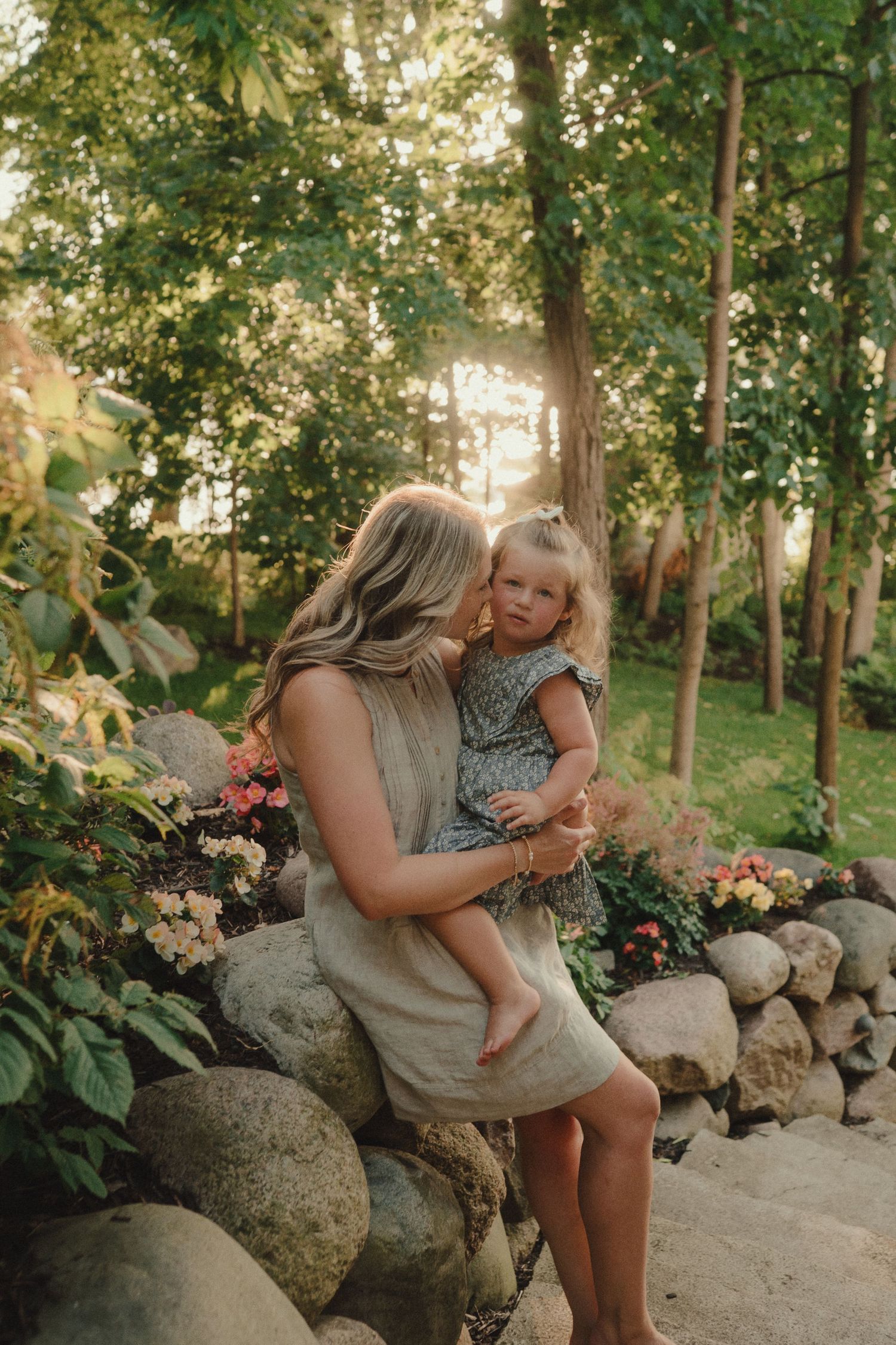 Mother kissing her toddler daughter while sitting on a stone wall surrounded by blooming garden flowers.