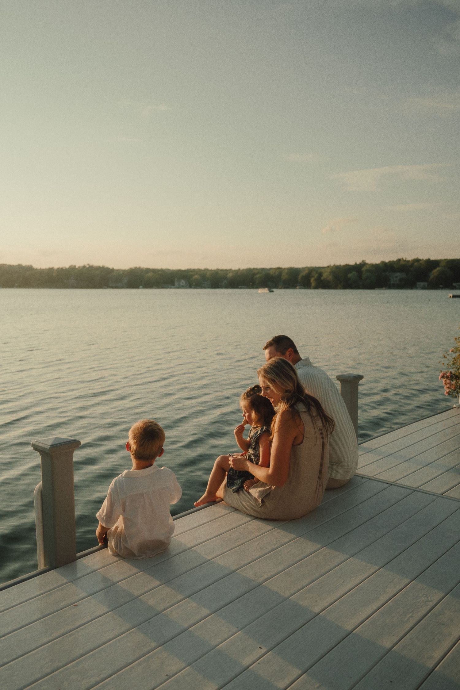 Family of four sitting on white wooden dock watching peaceful sunset over calm lake at dusk.