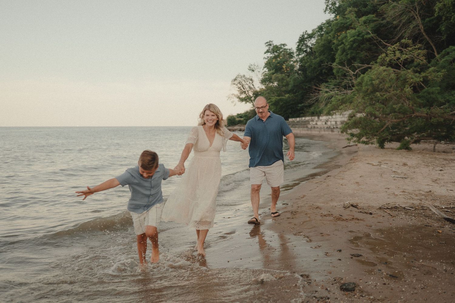 A joyful family of three walks along a sandy beach shoreline at sunset, splashing in the water together.