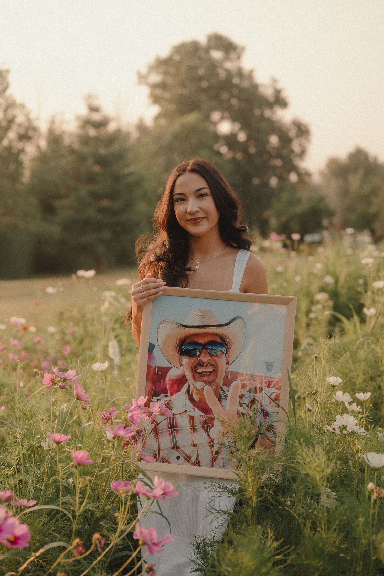 Young woman in white dress holding a painted portrait in a blooming wildflower field at golden hour.