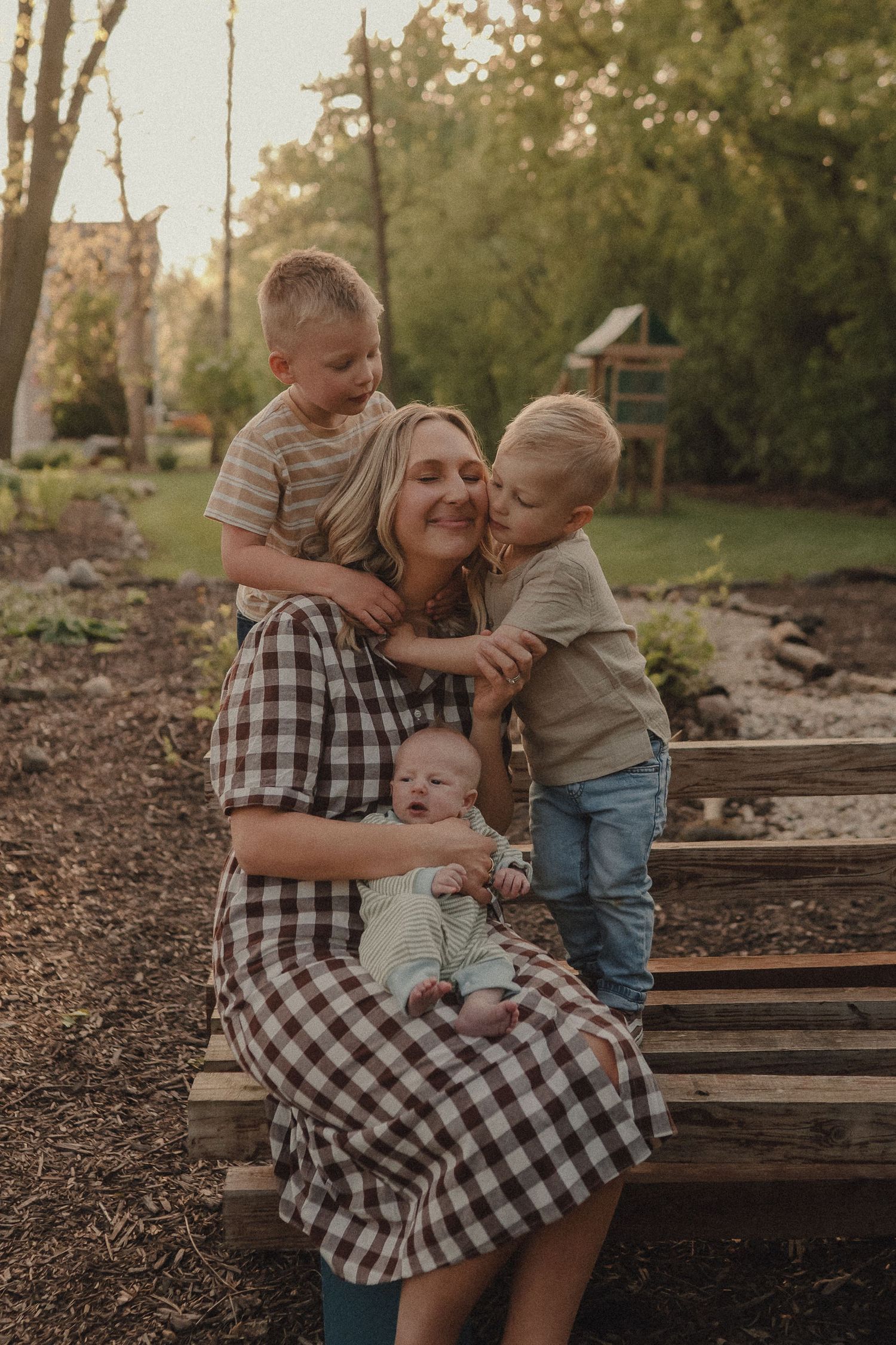A mother sits on a wooden bench holding her newborn while two young sons embrace her warmly.