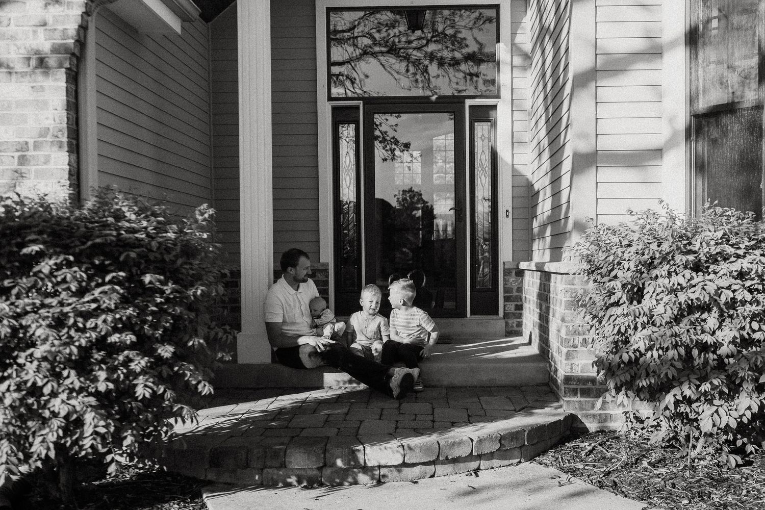 Black and white photo of a parent sitting with two young children on front porch steps of a suburban home.