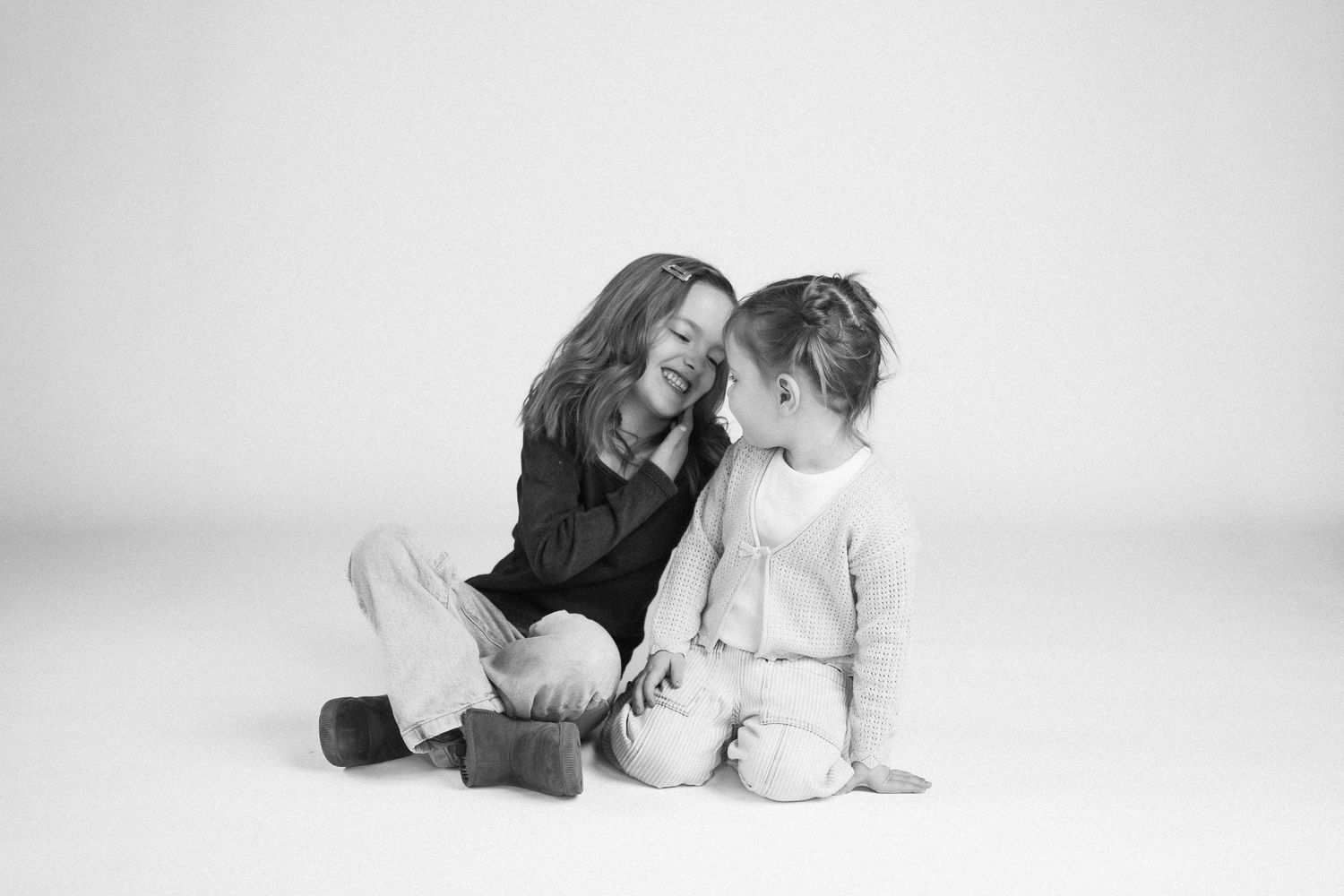 Black and white studio portrait of two people sitting close together on white background.