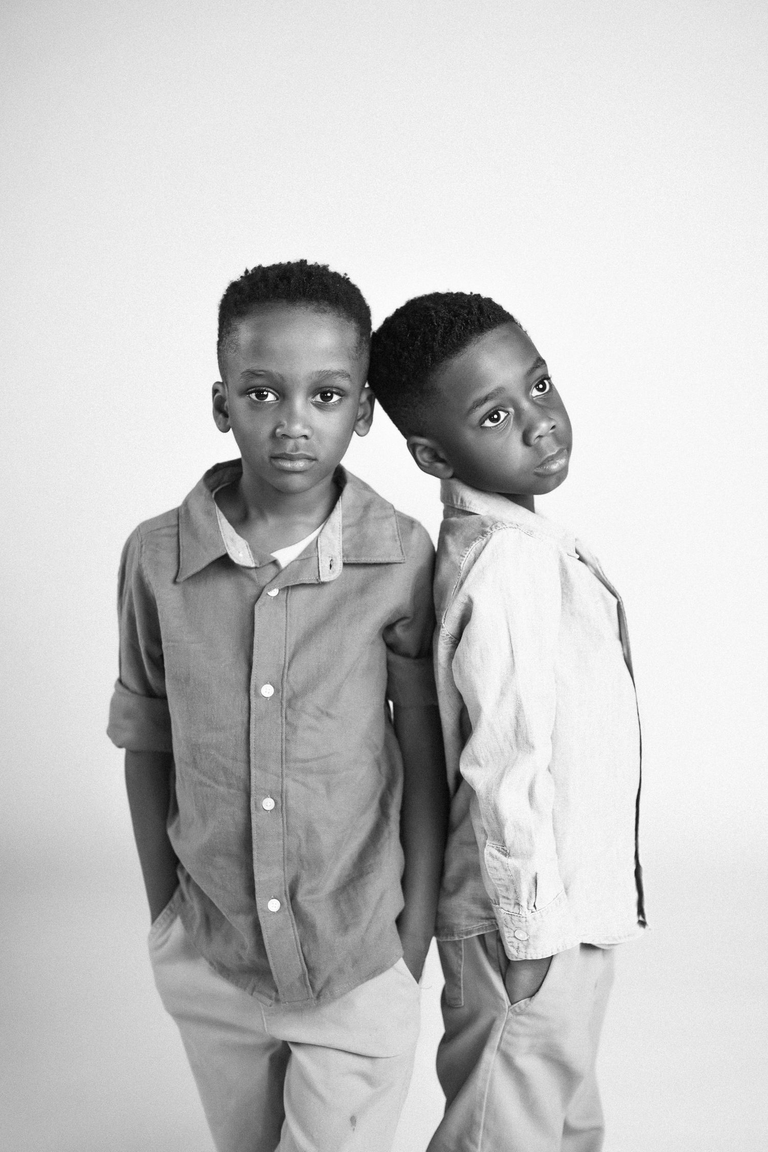Two children in button-down shirts pose back to back in a black and white studio portrait.