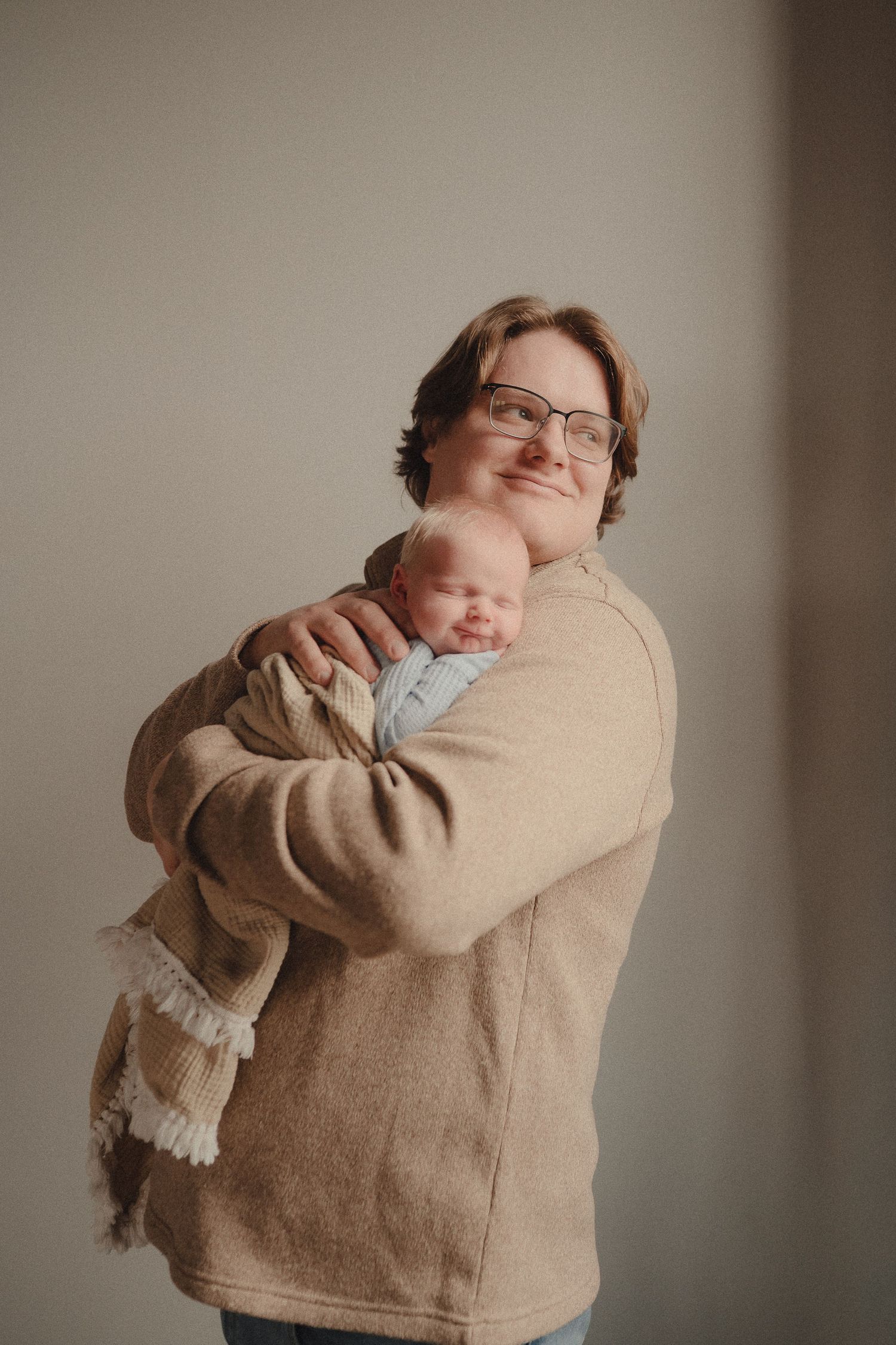 Smiling parent in beige sweater gently cradling a sleeping newborn baby against their chest.