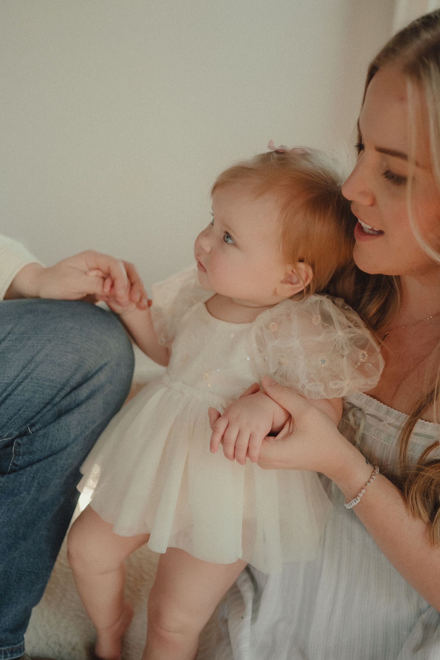 A blonde mother smiles warmly at her baby girl in a white dress in soft indoor light.