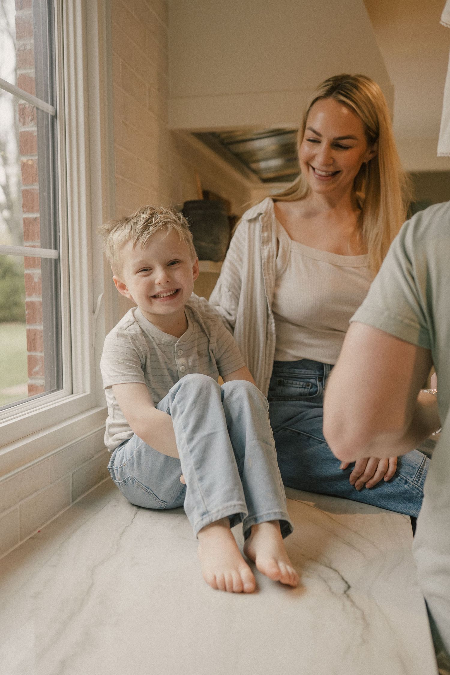 A smiling blonde mother and young barefoot boy sit together on a marble kitchen countertop by a window.