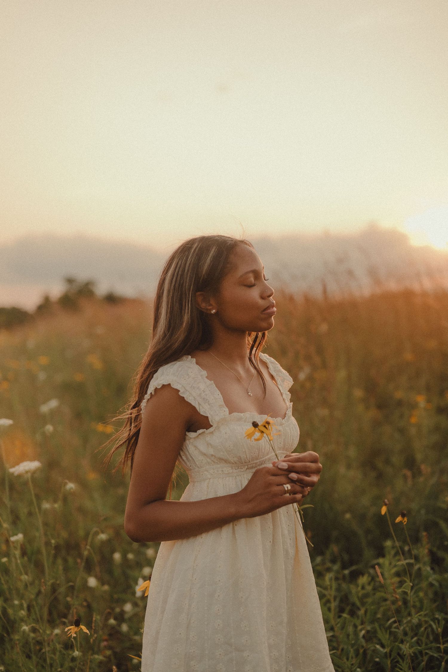 Young woman in white dress holds yellow wildflower with eyes closed at golden sunset.