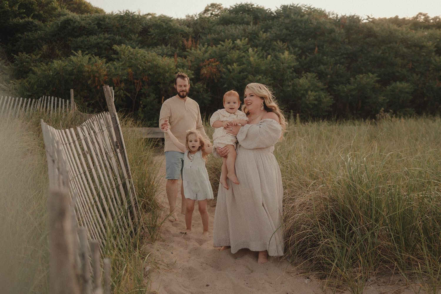 Family of four posing on sandy beach path surrounded by tall grass and wooden fence at golden hour.