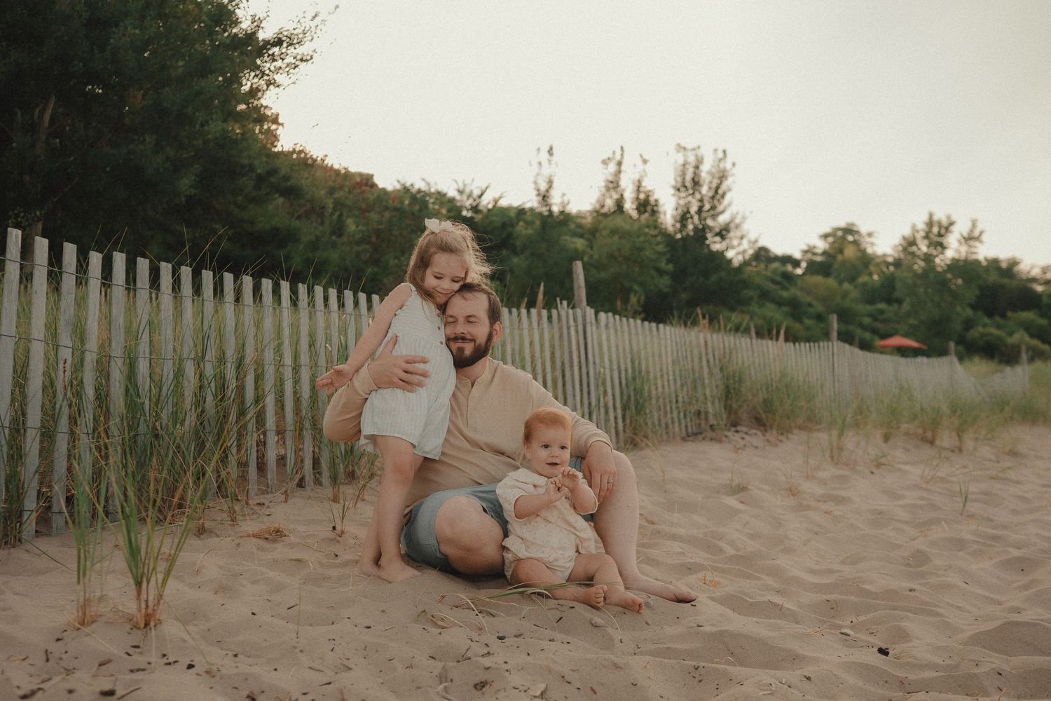 Father sitting on sand with two young children by beach fence.