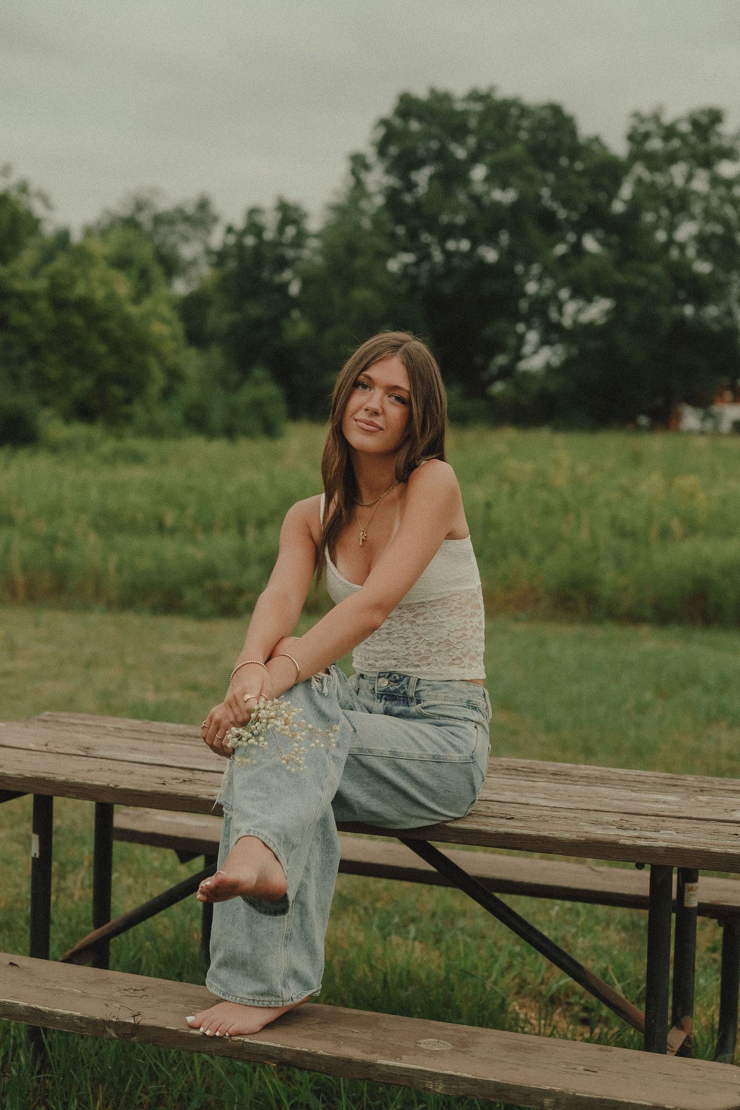 Young woman in white camisole and light blue jeans sitting barefoot on wooden bench in field.