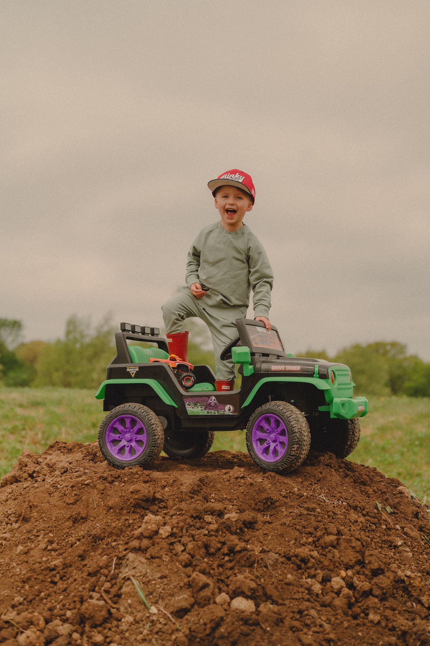 Excited boy in red cap stands on green ride-on Monster Jam Jeep on dirt hill.