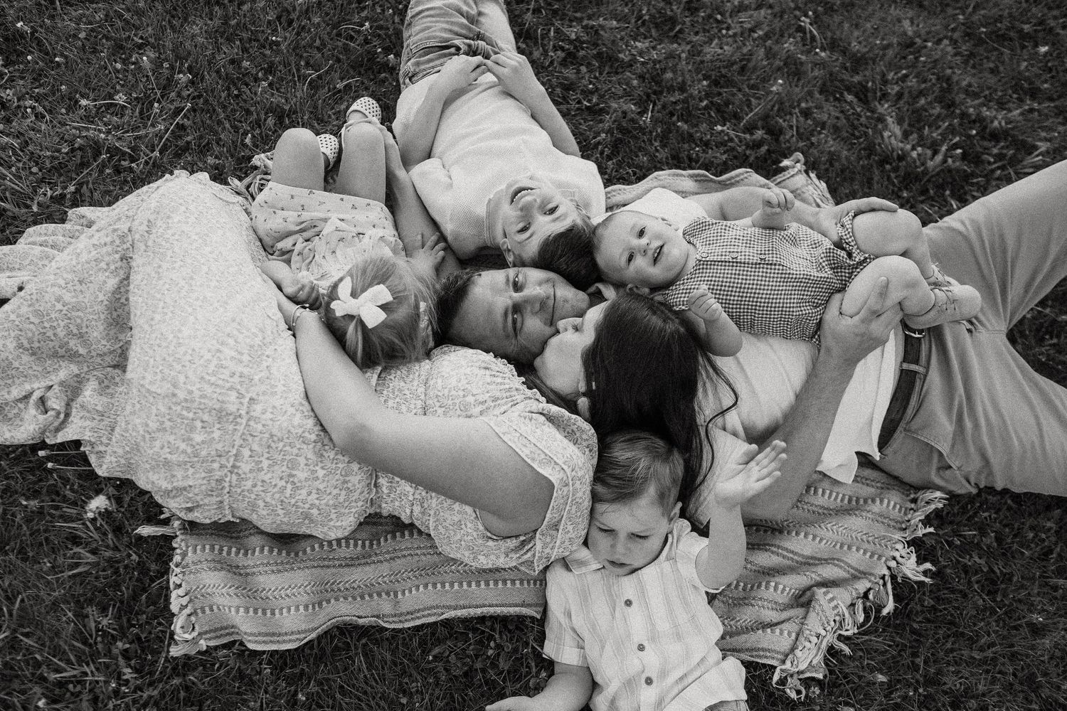 Black and white overhead photo of family lying on blanket in grass, smiling and cuddling together.
