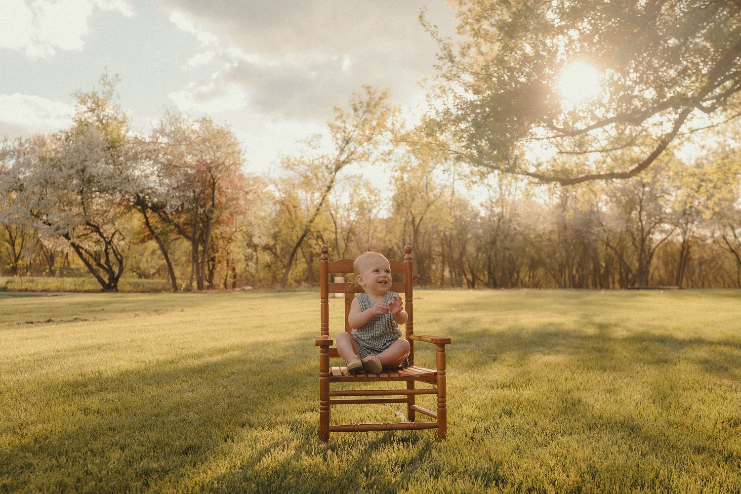 A baby sits on a small wooden chair in a sunlit park with golden autumn trees in the background.