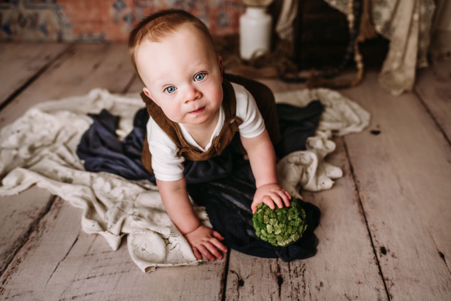 Crawling on wooden floor with decorative green prop and white fabric backdrop.
