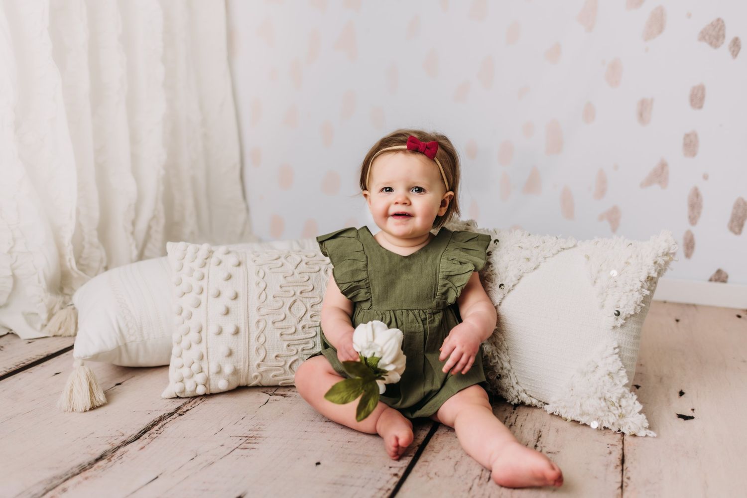 Baby in olive green romper sits among textured cream pillows.