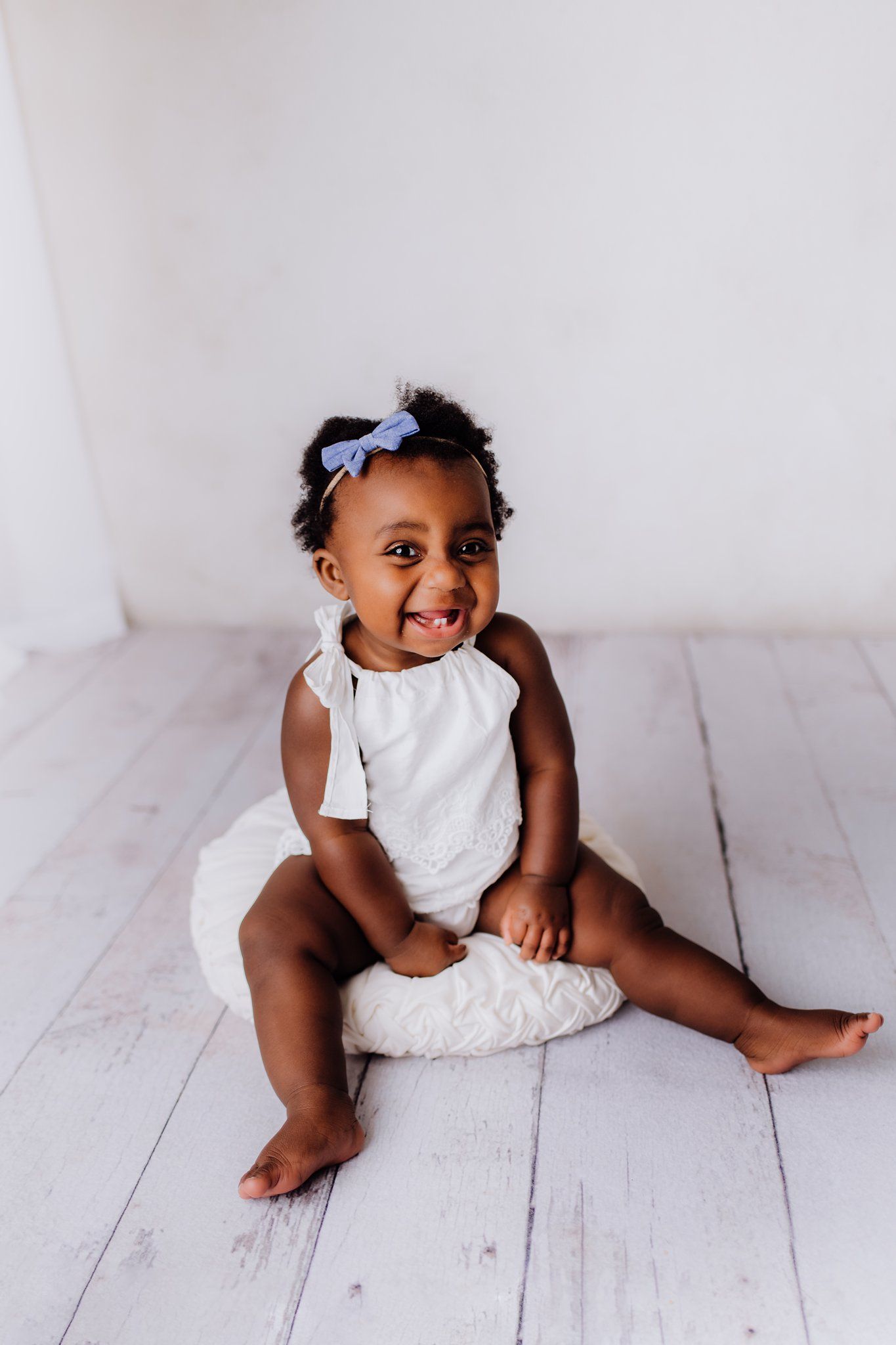 A happy toddler in a white outfit sits on a wooden floor with a bow accessory.