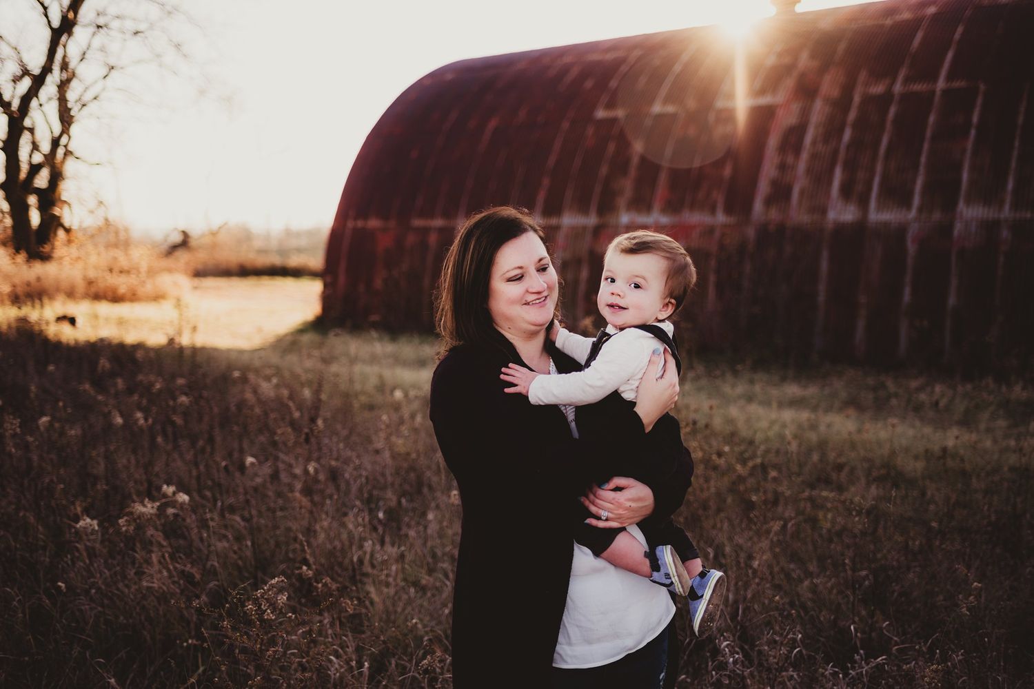 Mother and child share a tender moment at sunset in front of a rustic barn structure.
