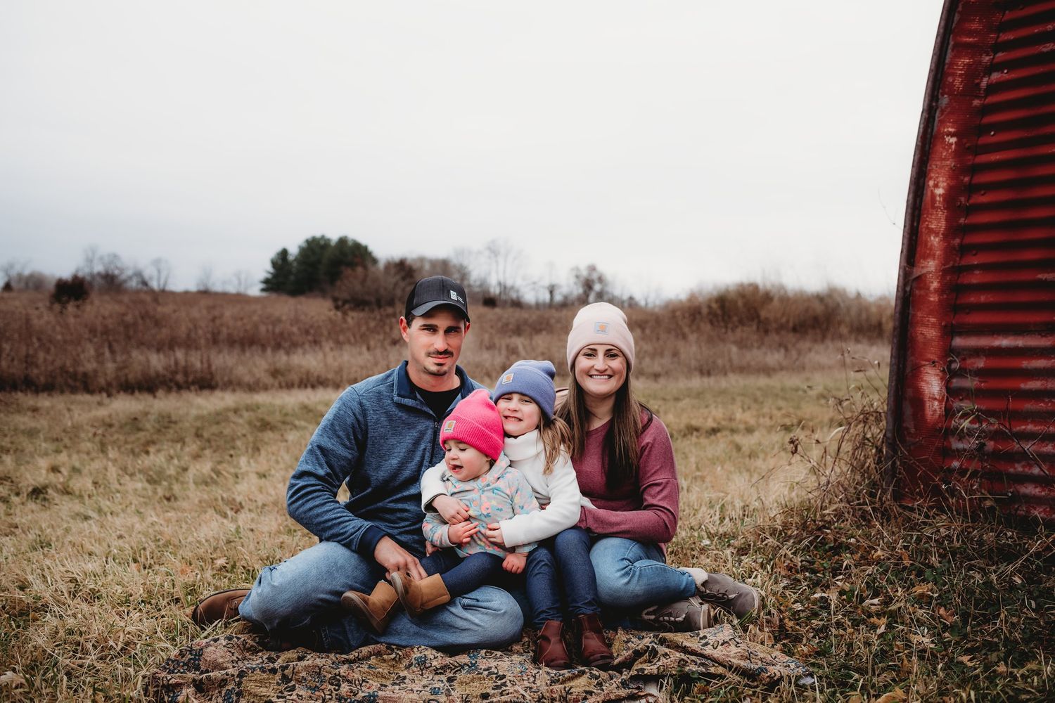 Family sitting together for casual outdoor portrait by rustic red barn.