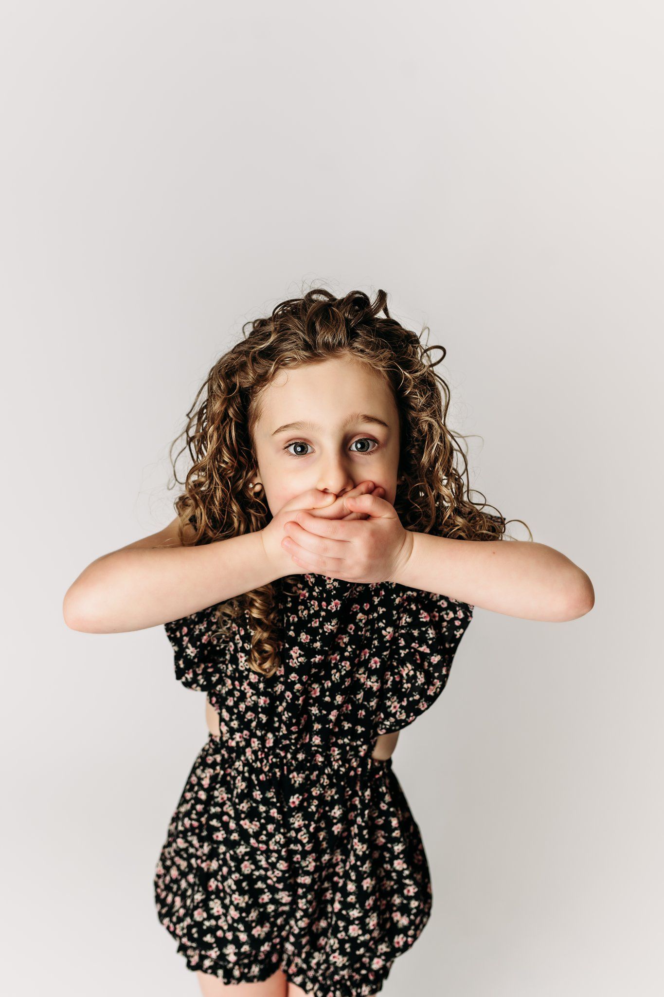 A child in a black floral dress covers their mouth with both hands on white background.