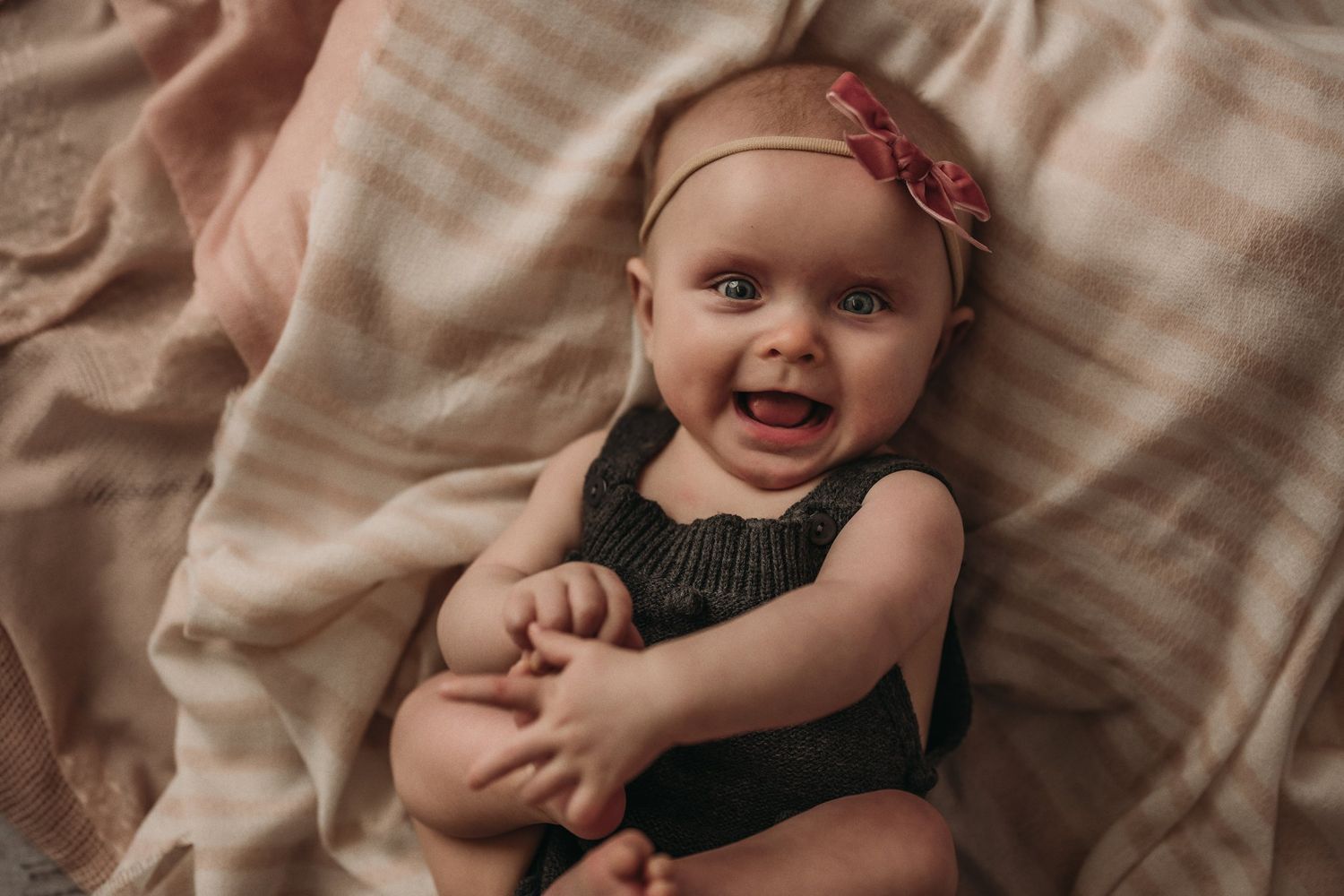 Baby smiling and hugging self while wearing dark romper on blush fabric.