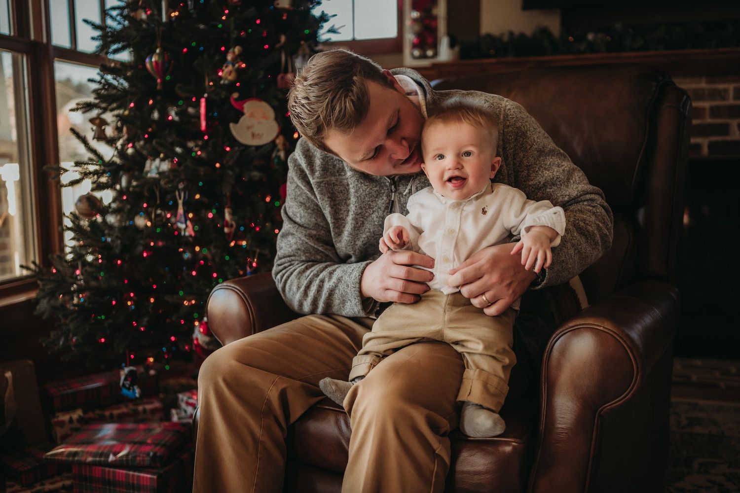 A tender holiday moment between parent and child sitting by a decorated Christmas tree near a window.