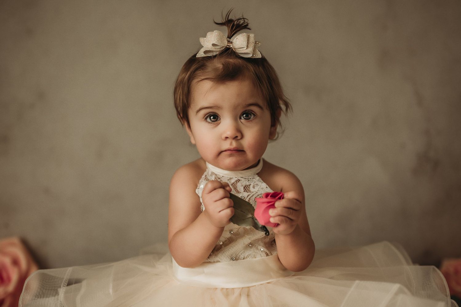 Little one in white tulle dress holding pink flower against neutral backdrop.