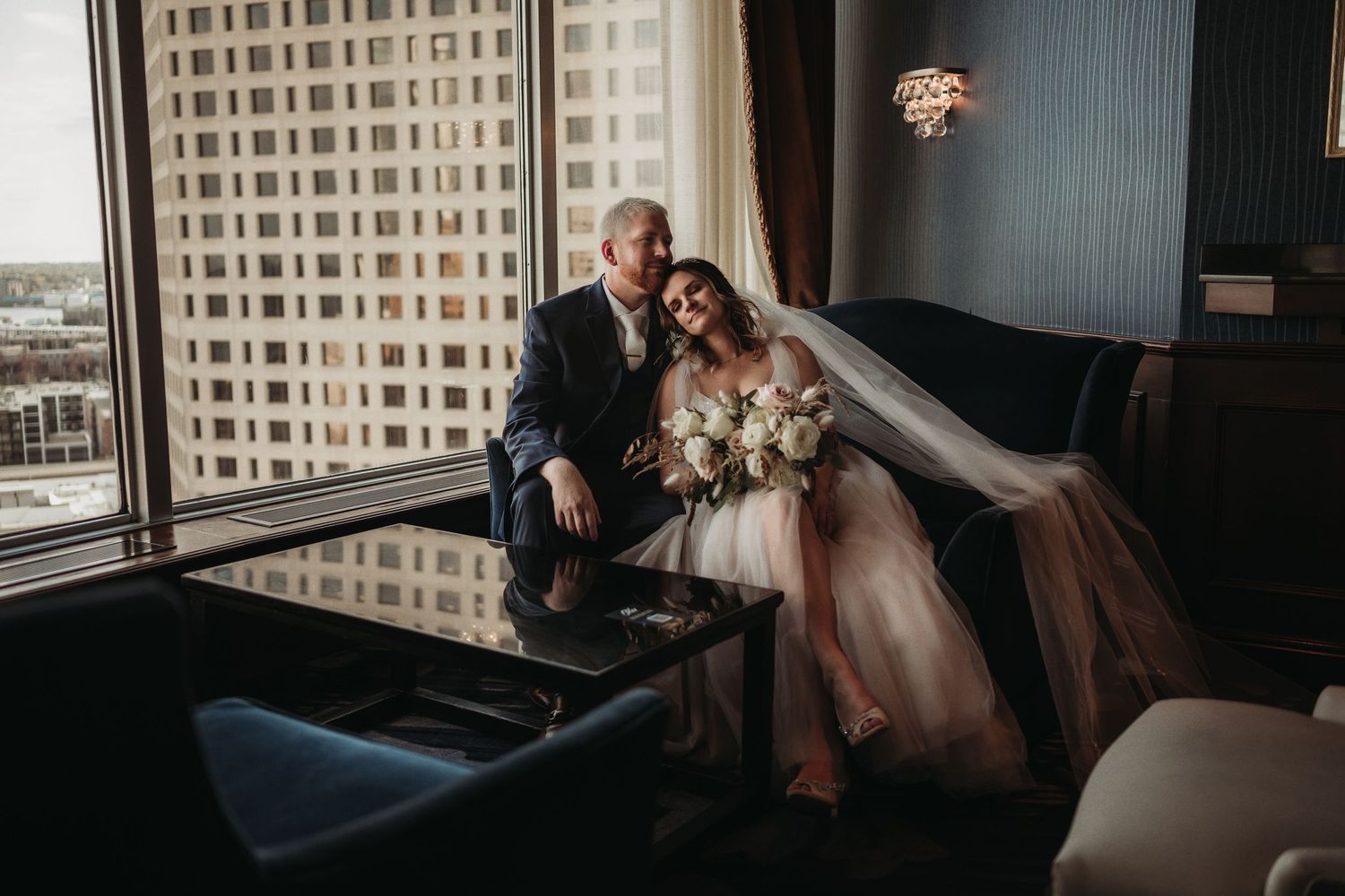 Couple in wedding attire sitting together in modern highrise room with city views.